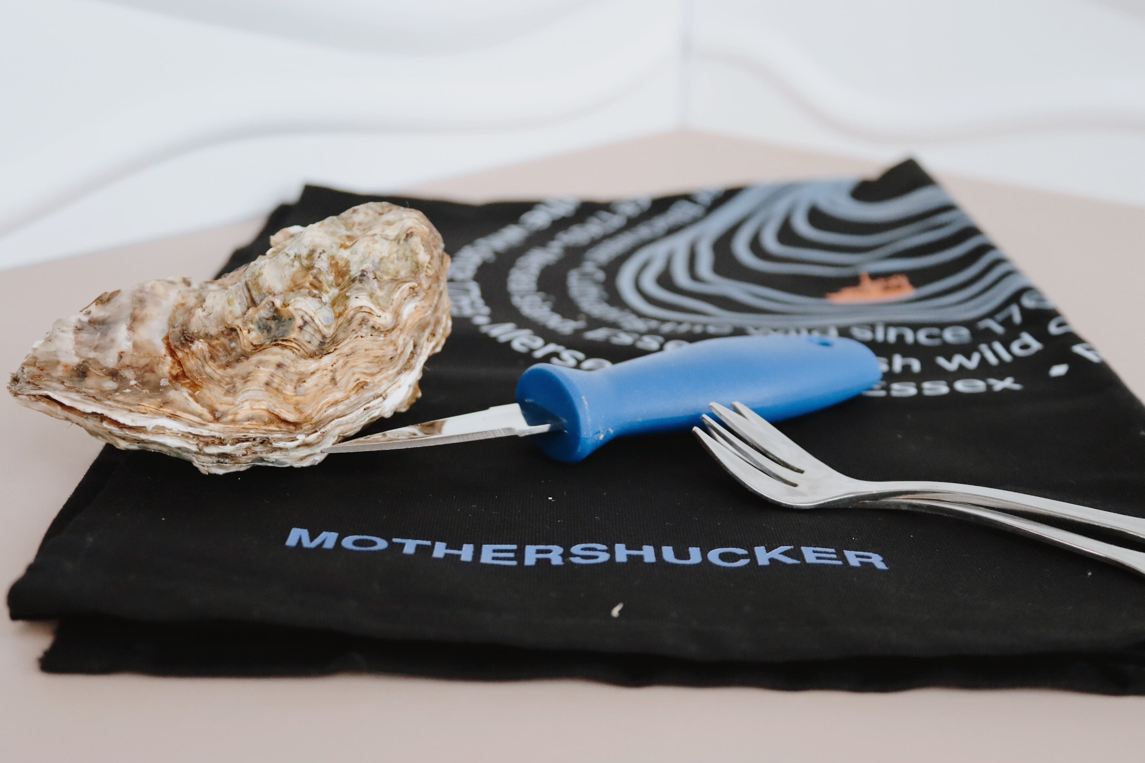 Mersea Oyster with a shucking knife on a black towel with 'Mothershucker' branding. Oyster forks. Shucking bundle. 