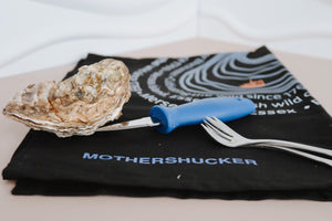 Mersea Oyster with a shucking knife on a black towel with 'Mothershucker' branding. Oyster forks. Shucking bundle. 