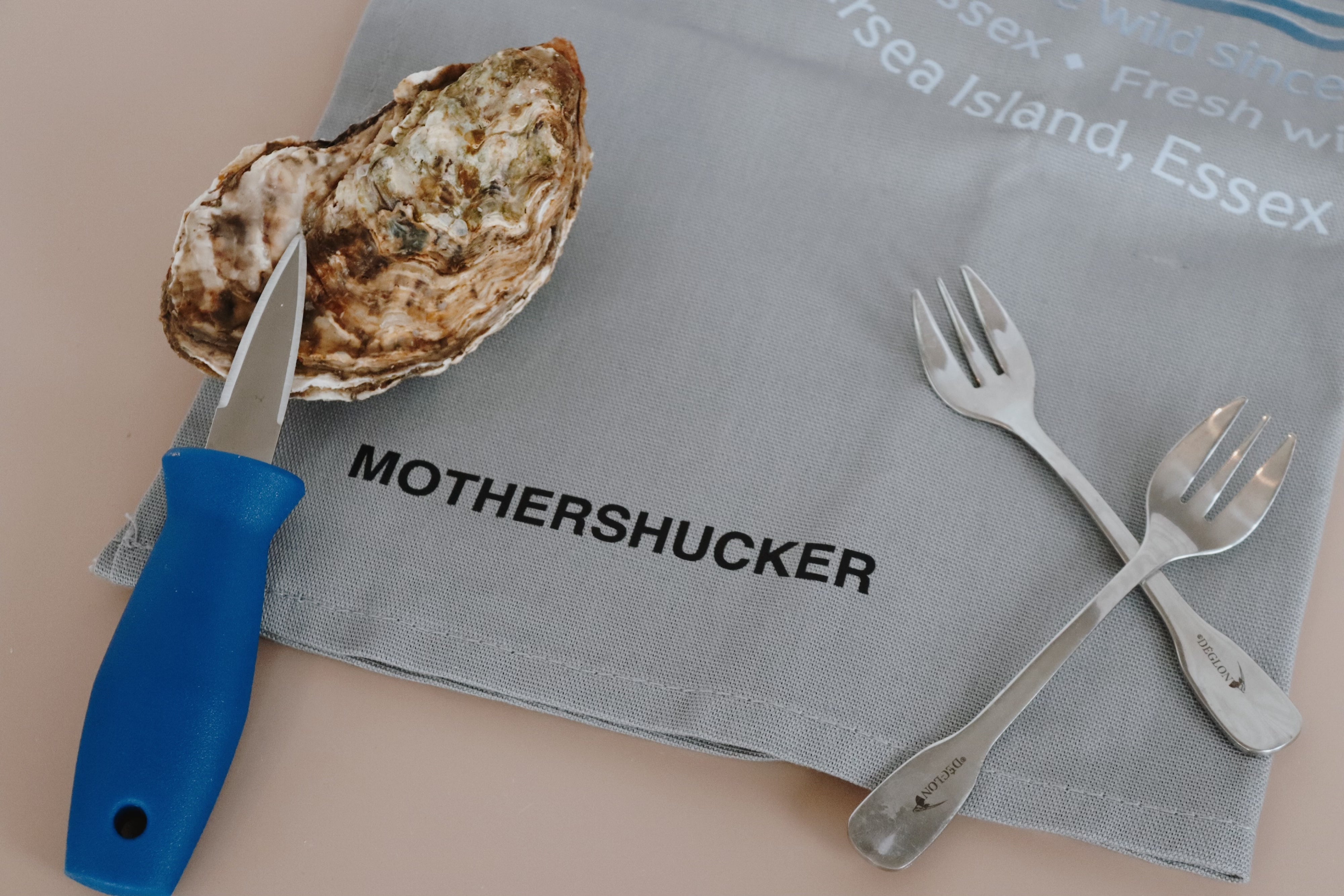 Oyster with a knife, two forks, and a napkin with 'MOTHERSHUCKER' text on a beige background. 