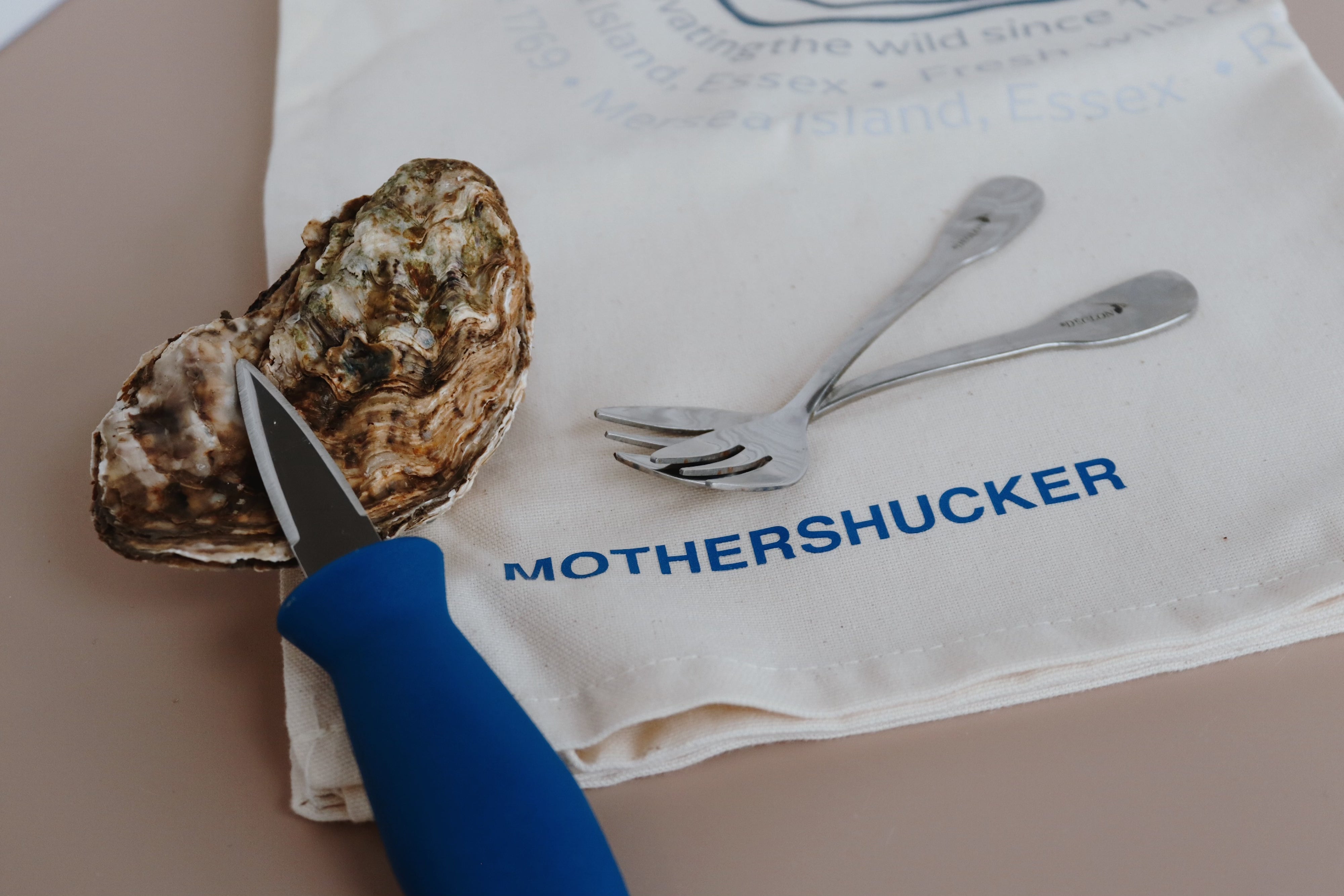 Oyster with a knife on a tea towel with 'MOTHERSHUCKER' branding.Natural colour. Oyster forks. 