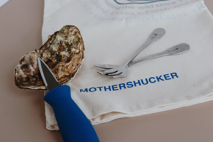 Oyster with a knife on a tea towel with 'MOTHERSHUCKER' branding.Natural colour. Oyster forks. 