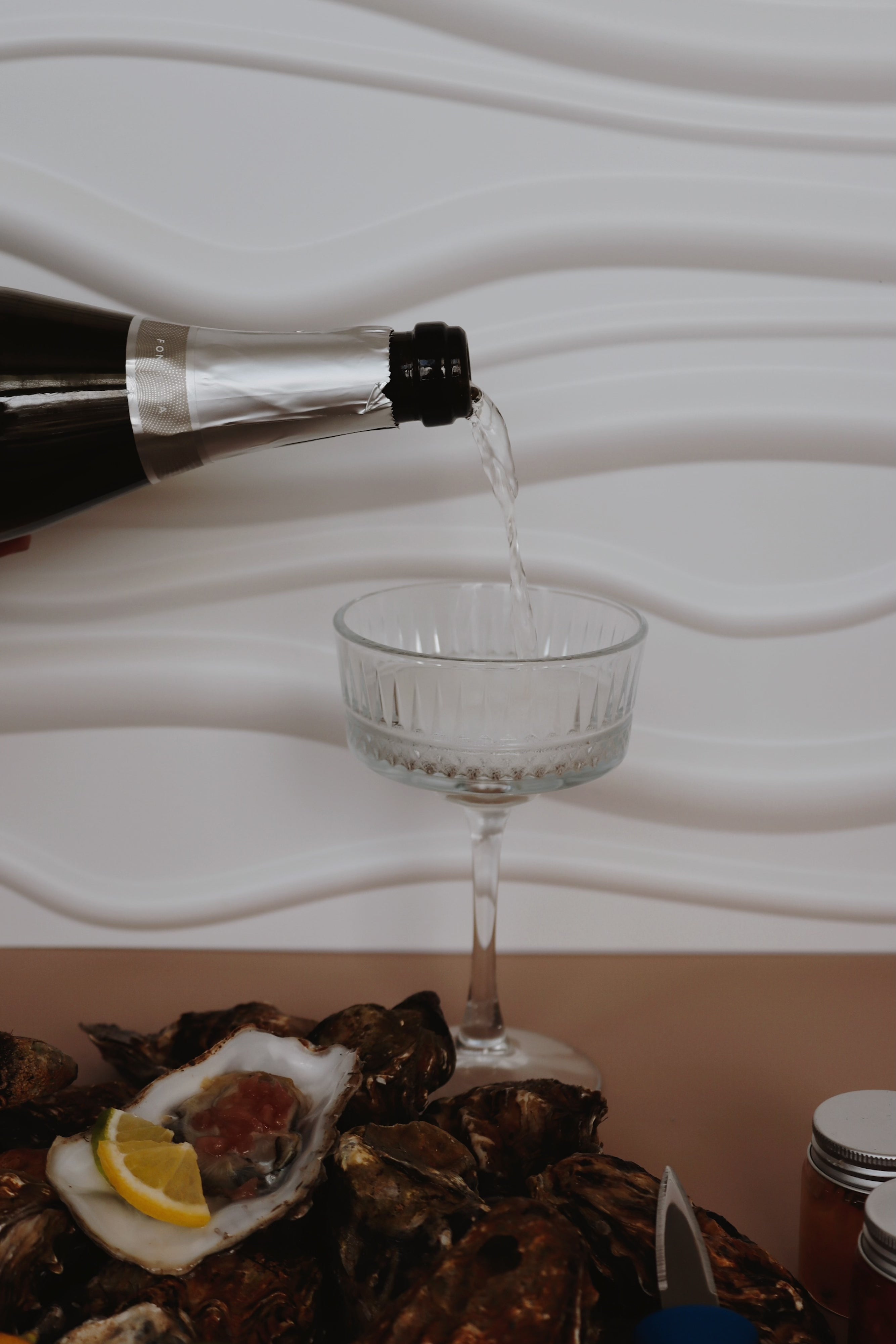 Bottle pouring Prosecco into a glass with Mersea rockl oysters and lemon on a white plate.