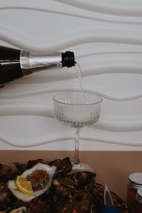 Bottle pouring Prosecco into a glass with Mersea rockl oysters and lemon on a white plate.