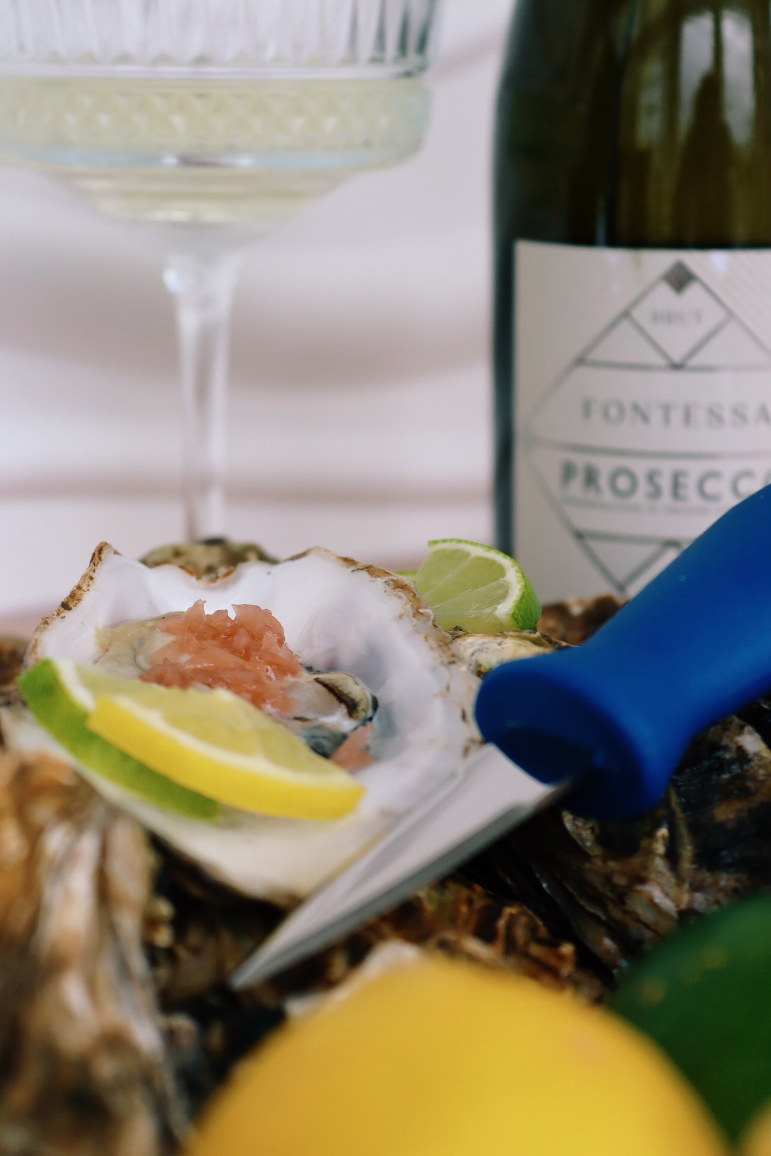 Mersea rock Oysters on a half shell with lemon wedges, a bottle of Fontessa Prosecco, and a glass of white wine. Square handled shucking knife. 