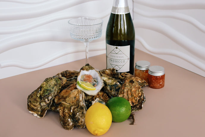 Mersea Oysters on a plate with a bottle of Prosecco and condiments on a beige surface.