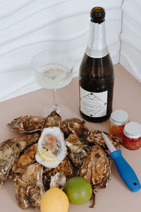 Oysters on a plate with a bottle of Prosecco, a glass, and condiments on a white background. Oyster shucking knife. 