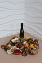 Load image into Gallery viewer, Mersea Oysters on the half shell with lemon wedges, a bottle of wine, and a small jar of sauce on a textured surface.