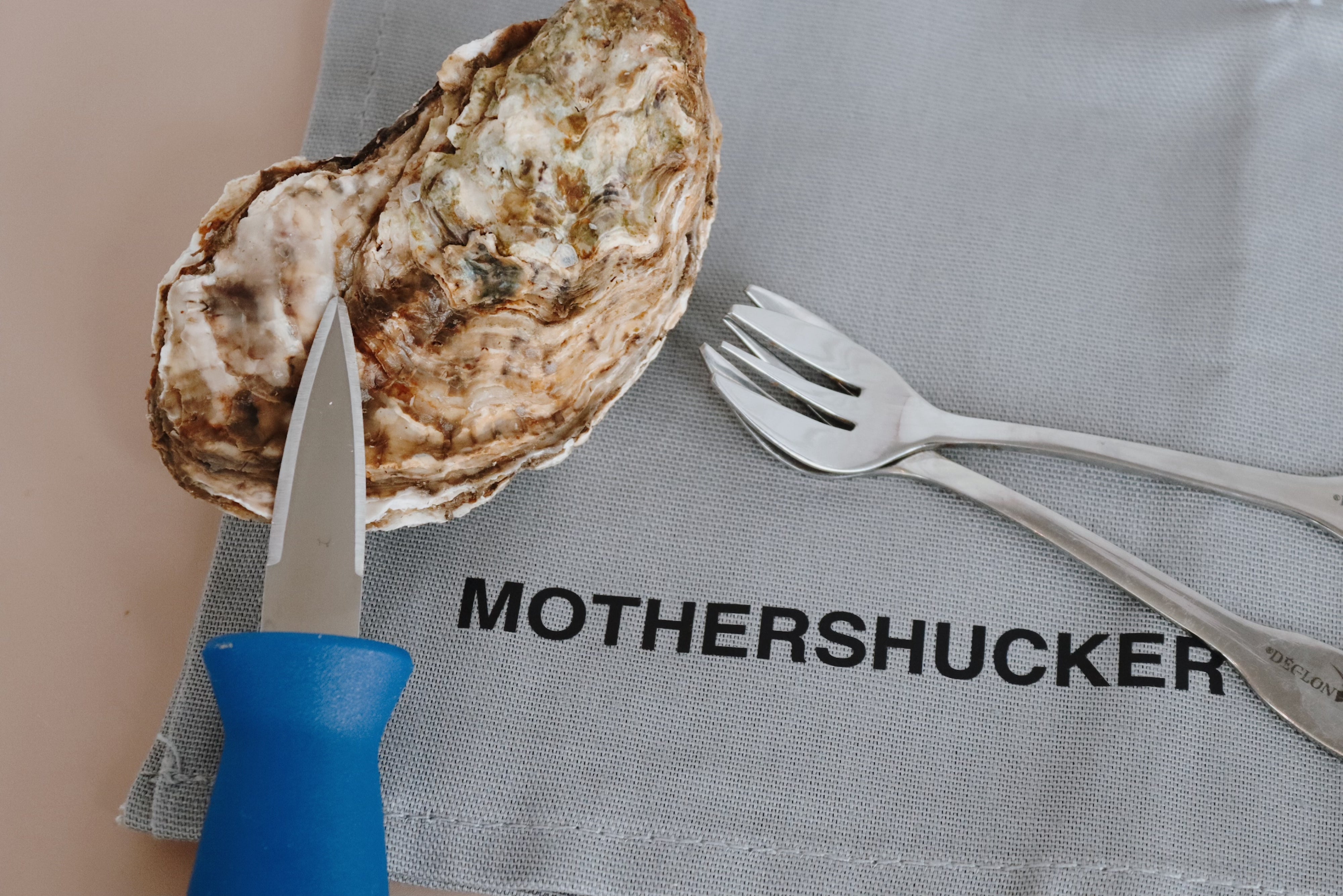Oyster with a knife, fork, and napkin with 'MOTHERSHUCKER' text on a neutral background