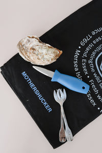 Oyster shucking tool, fork, and knife on a black towel with branding text.