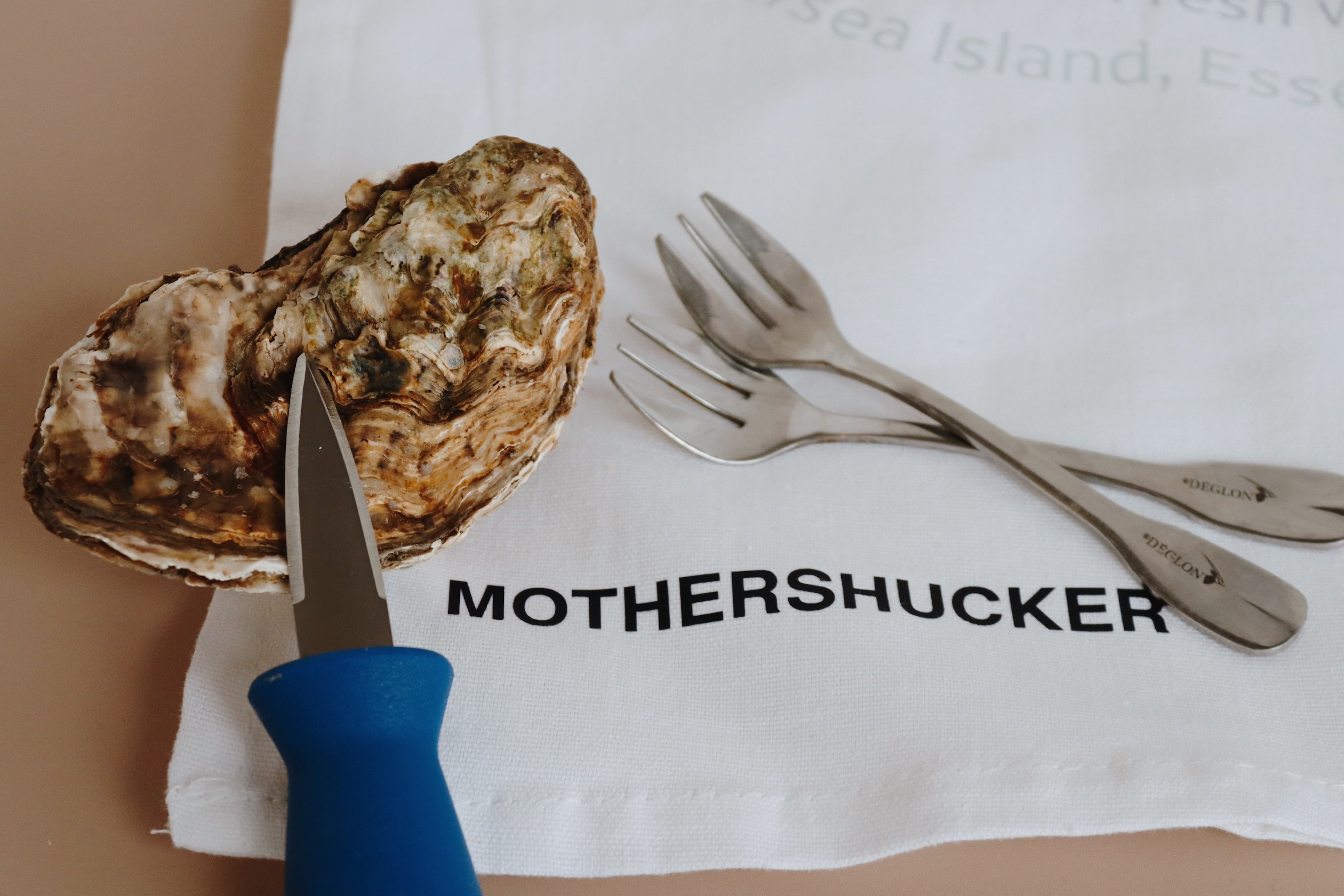 Oyster with a knife, two forks, and a white tea towel with 'MOTHERSHUCKER' text on a beige background.