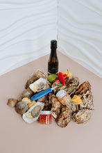 Load image into Gallery viewer, Assorted oysters on a half shell with garnishes and a bottle of wine on a beige surface.