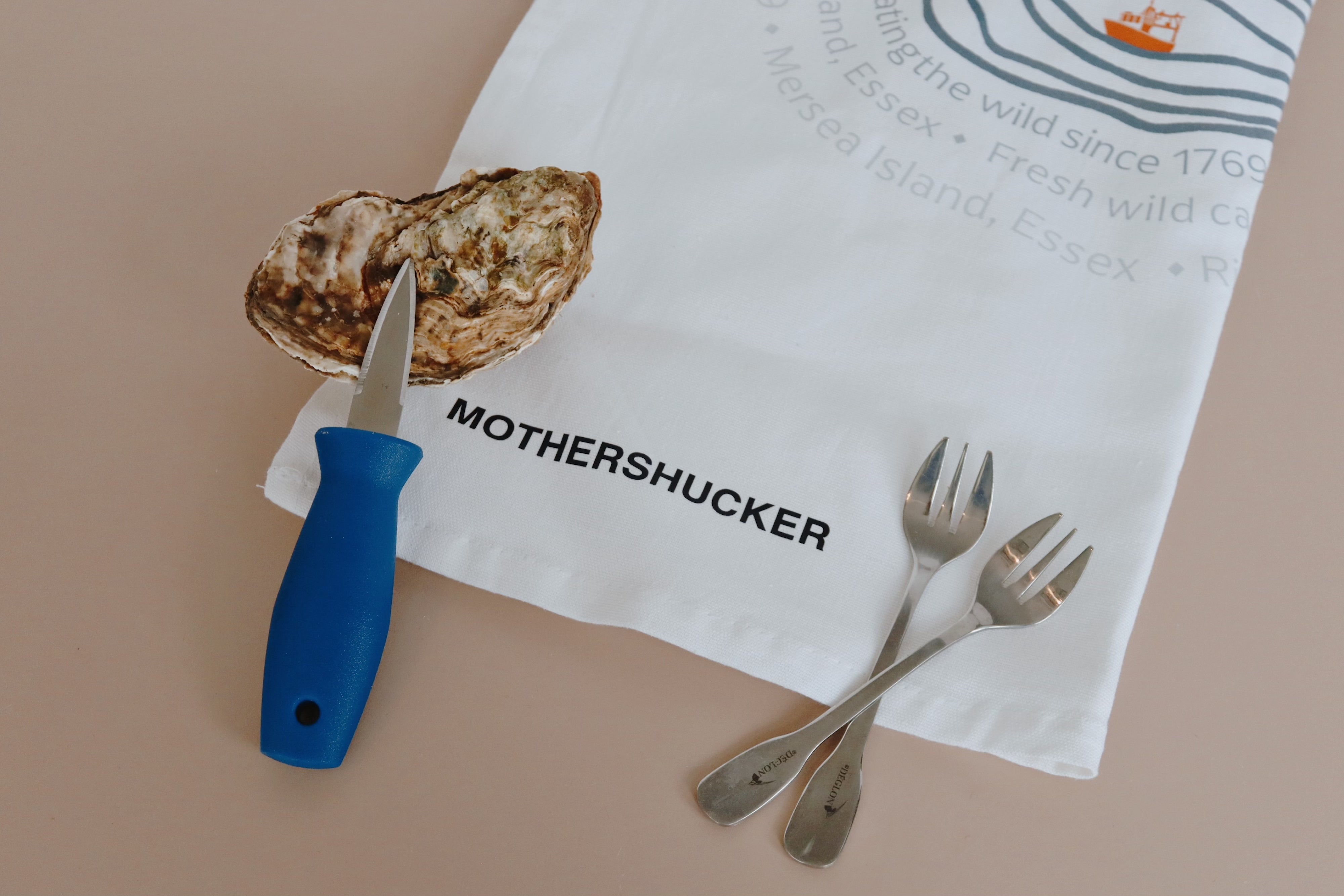 Mersea Oyster on a half shell with a knife and two forks on a 'Mothershucker' branded towel.